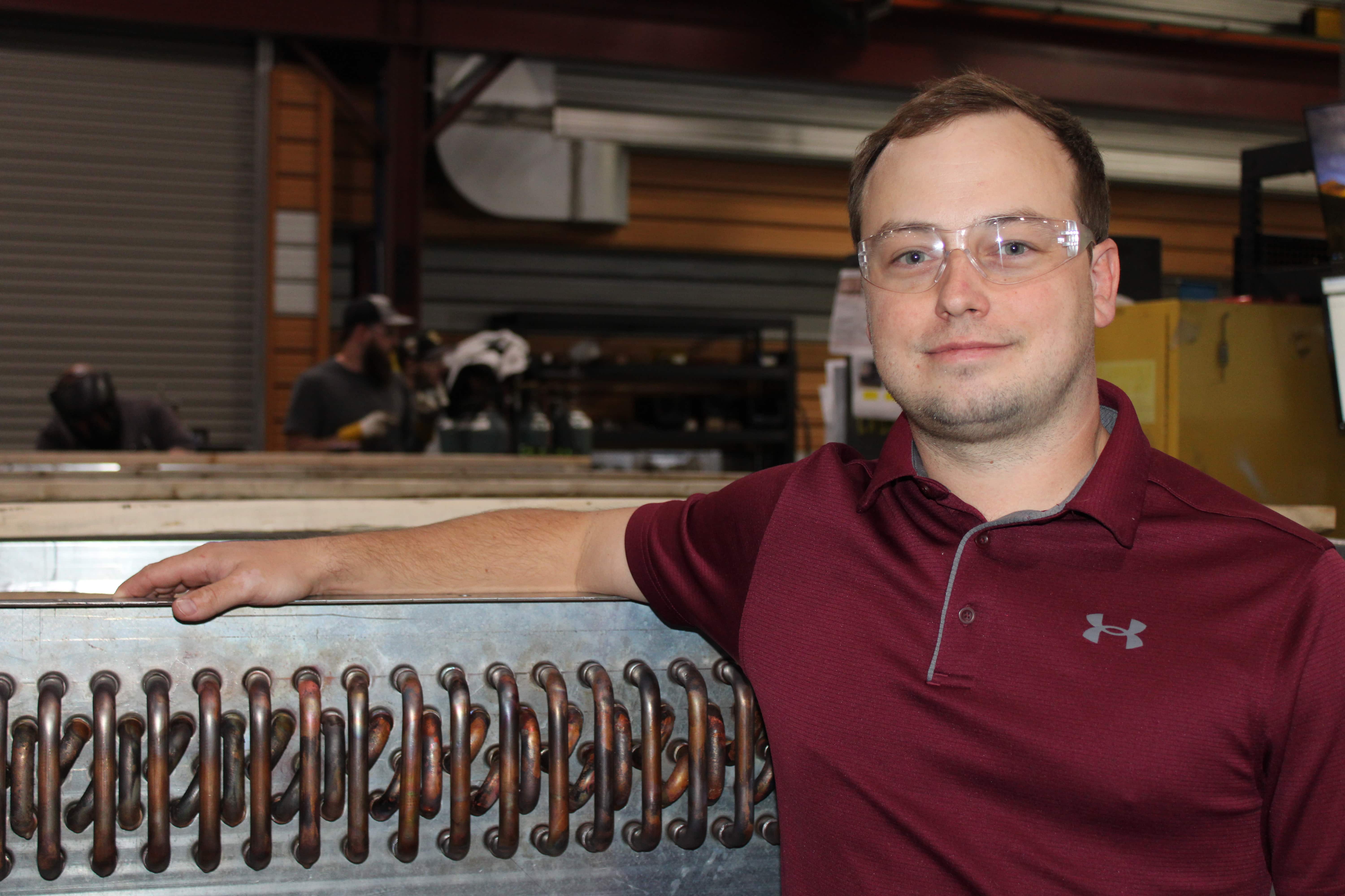 man in maroon shirt posing next to heat exchanger in manufacturing setting